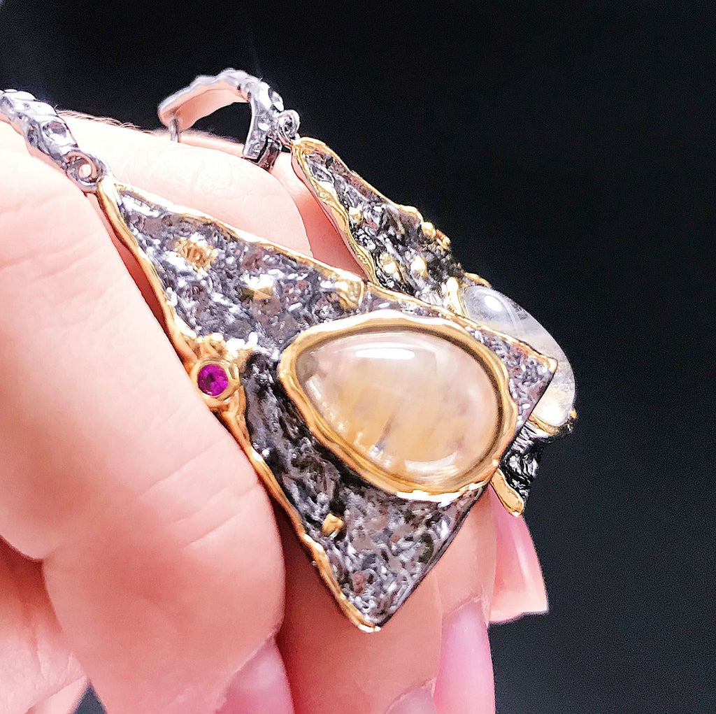 Gold and silver pendant with a central gemstone held by a hand against a dark background
