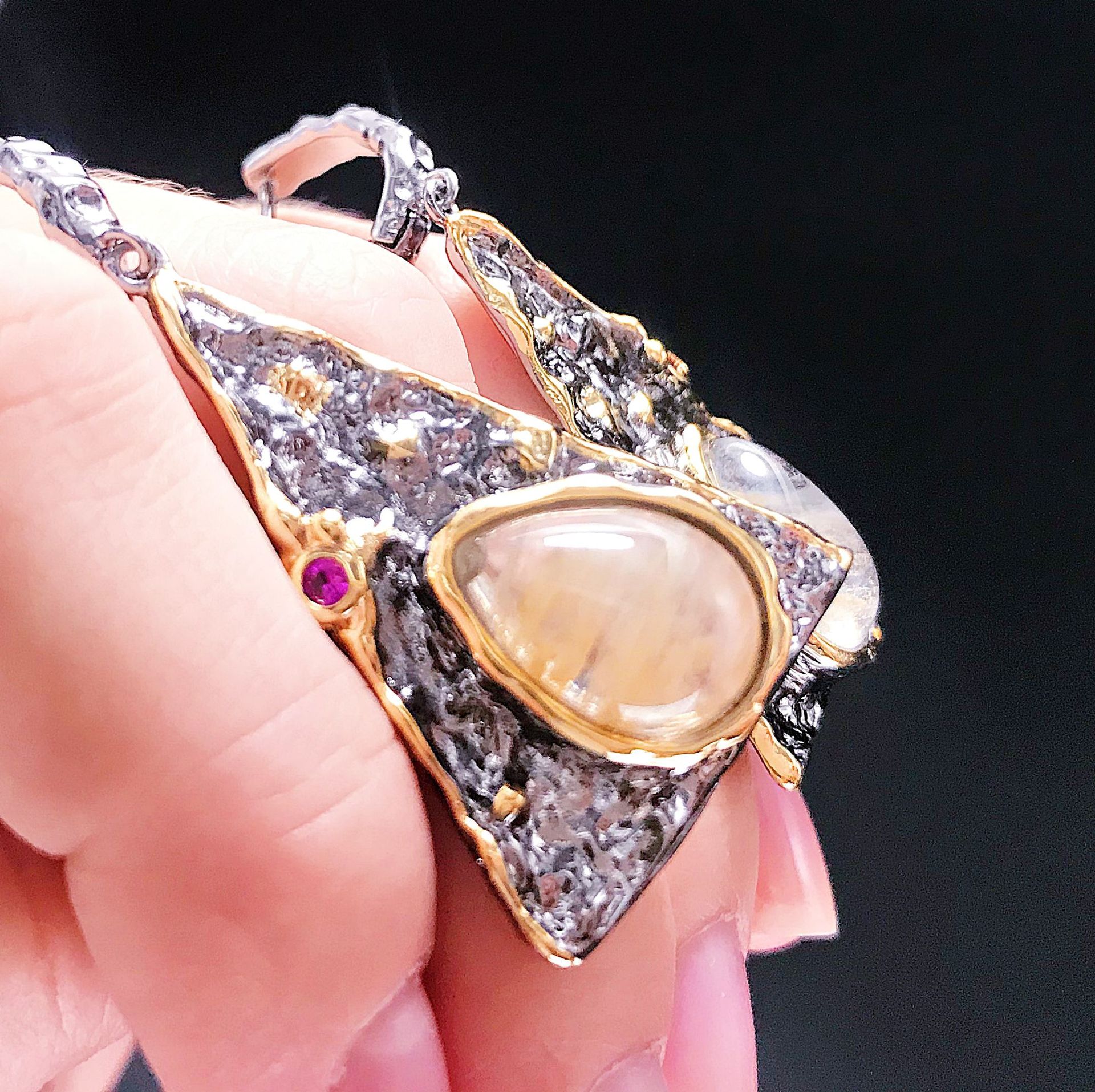 Gold and silver pendant with a central gemstone held by a hand against a dark background