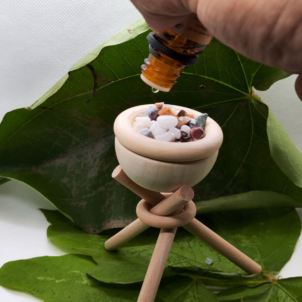 Small wooden bowl with stones on a stand, being dripped on by a hand holding a bottle over green leaves.