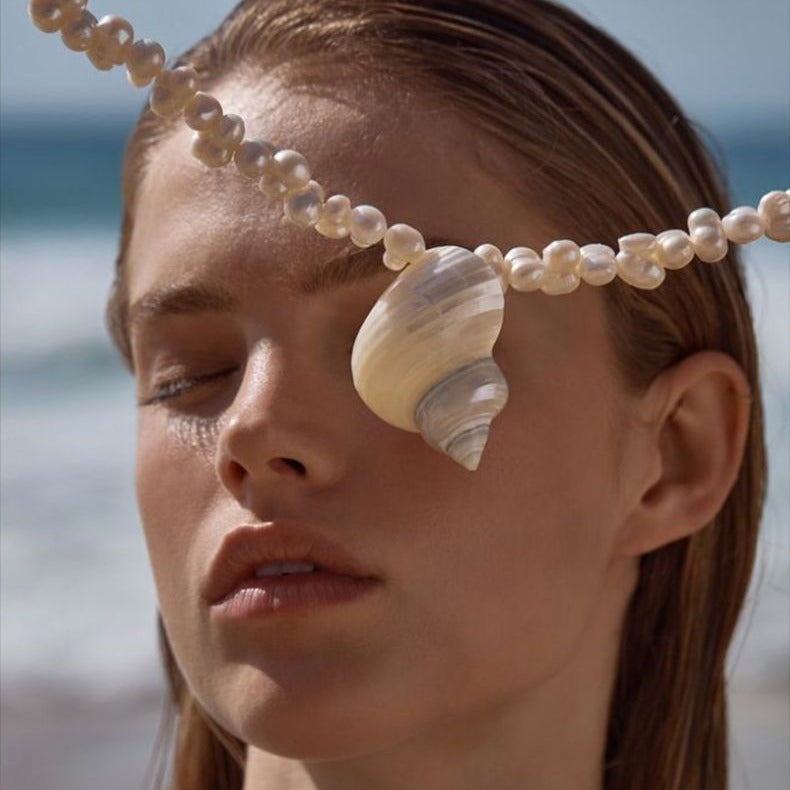 Woman with a shell on her face wearing a pearl headband against a beach background