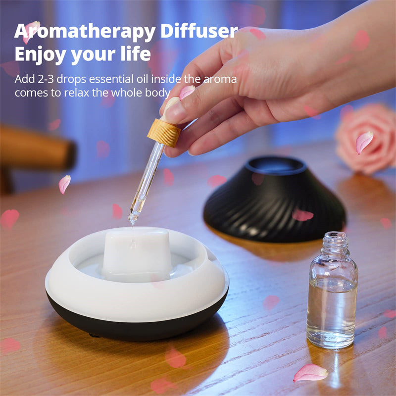 Aromatherapy diffuser with a hand adding essential oil, surrounded by essential oil bottle and flowers on a wooden surface.
