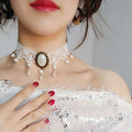 Close-up of a woman wearing a decorative lace choker necklace with pearls and a central stone, against a neutral background.