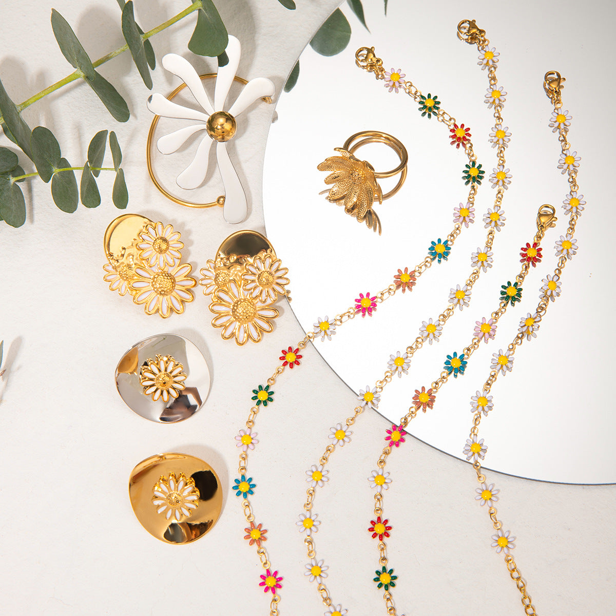 Gold jewelry including earrings, a ring, and a colorful floral necklace on a white surface with green leaves.