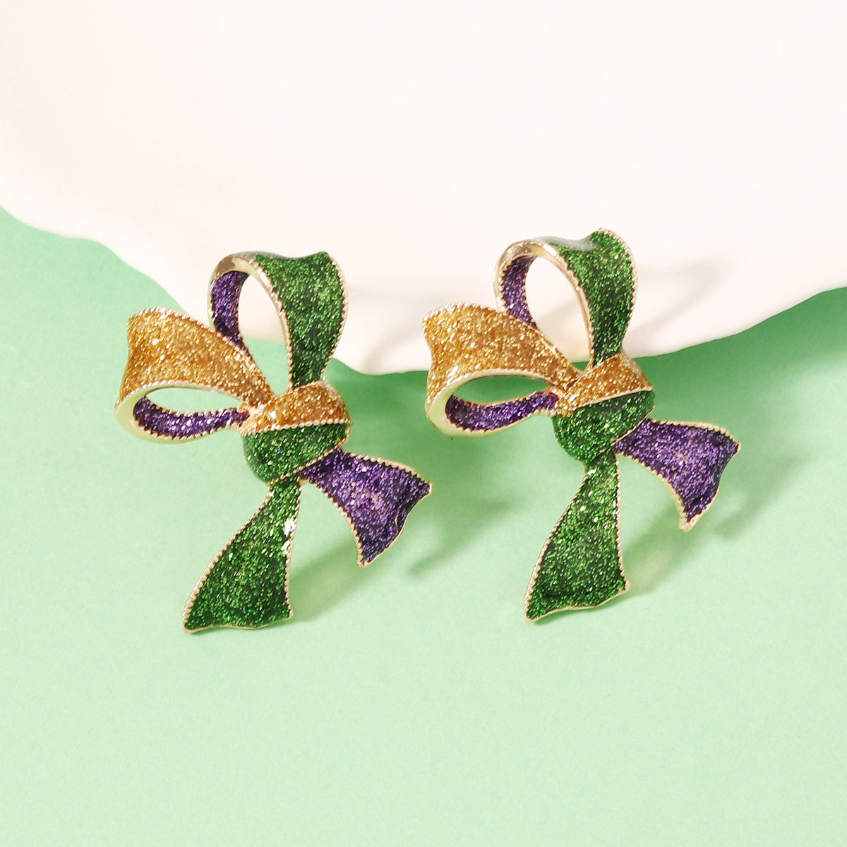 Pair of earrings shaped like green and gold ribbons with purple accents on a light green background